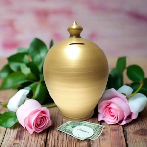 May include: A gold ceramic money box with a pointed lid. The money box is sitting on a wooden surface with pink roses and a pound note.