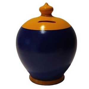Puede incluir: Una alcancía redonda de cerámica pintada a mano. La mitad superior está pintada de amarillo y la mitad inferior de azul marino. Tiene una ranura para monedas y un pequeño adorno decorativo en la parte superior. La base también es amarilla.