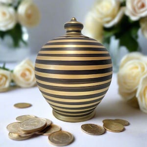 Op de afbeelding: Een bolvormige spaarpot met horizontale zwarte en metallic gouden strepen en een kleine knop aan de bovenkant. Het object heeft een gleuf voor munten en is omringd door verschillende verspreide goudkleurige munten op een plat oppervlak.
