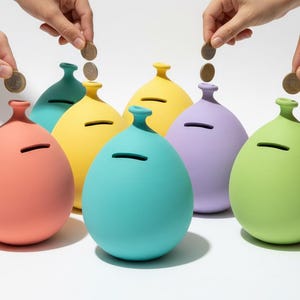Puede incluir: Una colección de huchas de colores en forma de globo, en tonos coral, turquesa, amarillo, morado y verde. Se están introduciendo monedas en las ranuras de las huchas. Las huchas están sobre una superficie blanca.