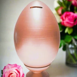 Könnte beinhalten: Eine eiförmige Spardose mit metallischem Roségold-Finish. Das Objekt verfügt über einen horizontalen Münzschlitz an der Oberseite und ruht auf einem runden Sockel. Die Oberfläche hat eine glatte, reflektierende Textur, die von einem dunkleren Kupferton oben zu einem helleren, blassrosa Farbton nach unten hin übergeht.