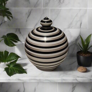 Può includere: Un vaso decorativo in ceramica con coperchio. Il vaso è sferico con strisce orizzontali nere, beige e bianco sporco. Il coperchio è una versione più piccola del vaso, con una parte superiore a punta. Il vaso è esposto su una superficie di marmo bianco.
