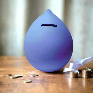 Pode incluir: Um cofrinho em forma de gota, azul-pervinca, com uma ranhura para moedas. Várias moedas estão espalhadas na superfície de madeira, com duas pilhas de moedas e uma fita branca por perto. O cofrinho é um item decorativo para poupar dinheiro.