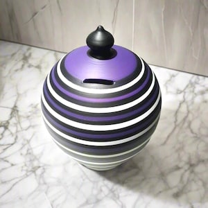 May include: A round ceramic bank with a black, white, and purple striped pattern. The top is solid purple with a slot for inserting coins. A small, black, rounded knob sits atop the lid. The bank is on a marble surface.