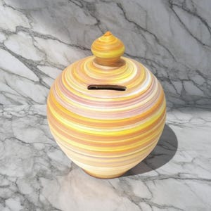 Può includere: Un salvadanaio in ceramica con un design a spirale nei toni del giallo, arancione e bianco. La banca ha una fessura per le monete e un coperchio rimovibile. Il salvadanaio rotondo è un oggetto decorativo.