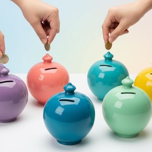 Pode incluir: Uma coleção de mealheiros de cerâmica redondos e coloridos em tons de roxo, coral, verde-azulado, verde, azul e amarelo. Mãos depositam moedas nas ranhuras superiores. Os mealheiros têm um acabamento brilhante.