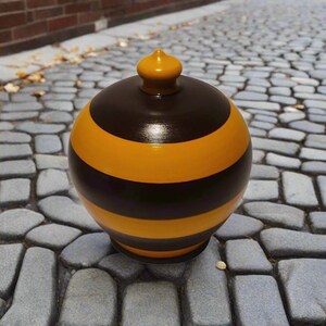 Peut inclure: Un pot en céramique sphérique avec une finition brillante, présentant des rayures horizontales alternées de couleur brun foncé et jaune moutarde. Le pot possède un couvercle assorti jaune moutarde avec un petit bouton arrondi.
