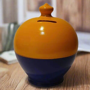 Può includere: Un salvadanaio rotondo bicolore. La metà superiore è arancione lucido e la metà inferiore è blu scuro. Ha una fessura per inserire le monete e un piccolo ornamento decorativo sulla parte superiore. Il salvadanaio è su una superficie di legno.