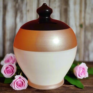 May include: A ceramic money bank with a rounded shape, featuring a color scheme of white, bronze, and dark brown. The bank has a slot on top and is decorated with pink roses. The bank is designed for saving money.