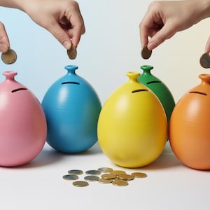 Puede incluir: Cinco alcancías de cerámica de colores rosa, azul, amarillo, verde y naranja están dispuestas sobre una superficie blanca. Manos introducen monedas en las ranuras de las alcancías. Un montón de monedas está delante.
