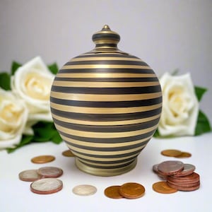 May include: A round, black and gold striped ceramic piggy bank with a matching lid. The bank is surrounded by loose coins and white roses. The bank is designed to hold savings.