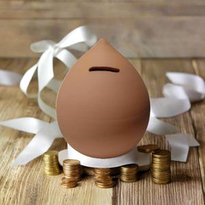 May include: A brown, teardrop-shaped piggy bank with a slot for inserting coins. Several stacks of coins are arranged in front of the bank. White ribbon and a bow are in the background, set against a wooden surface.