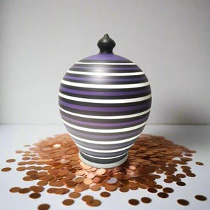 Peut inclure: Un pot décoratif en céramique avec un motif rayé noir, violet et blanc. Le pot a un bouton noir sur le dessus et repose sur une surface recouverte de confettis couleur cuivre. Un objet de décoration intérieure unique.
