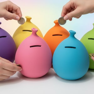 Puede incluir: Seis huchas de colores en forma de huevo, en morado, amarillo, rosa, naranja, azul y verde. Cada una tiene una ranura para monedas y unas manos están añadiendo monedas. Las huchas están sobre una superficie blanca.