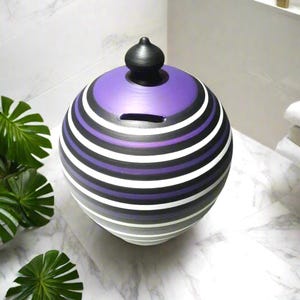 Puede incluir: Un tarro de cerámica redondo y decorativo con tapa, con rayas horizontales en morado, negro, blanco y gris. El tarro tiene una pequeña ranura y un pomo negro en la parte superior. Está sobre una superficie de mármol.