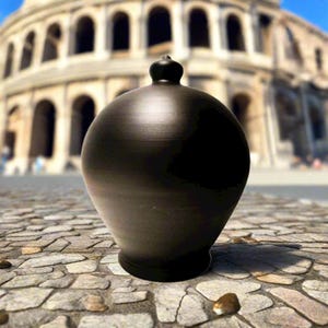 May include: A dark, round ceramic vase with a small, decorative top. The vase has a smooth, matte finish and is placed on a cobbled surface. The background shows a blurred view of a large, ancient Roman structure.