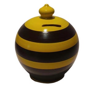 May include: A spherical ceramic piggy bank featuring horizontal stripes in alternating yellow and dark brown colors. The top of the bank has a small, rounded yellow lid with a knob handle and a horizontal coin slot positioned just below the lid.
