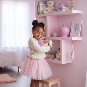 May include: A young girl in a white sweater and pink polka dot tutu holds a pink object in a pink room. A pink shelf displays books, a teddy bear, and a framed picture. A ballet theme is present with ballet shoes hanging from the shelf.