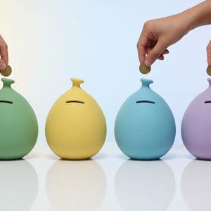 May include: Four colorful piggy banks in green, yellow, blue, and purple, each with a coin slot. Hands are dropping coins into the slots. The banks have a rounded shape with a small neck.