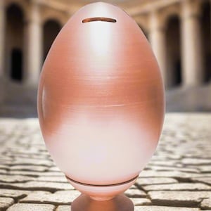 Può includere: Un salvadanaio a forma di uovo color rame con una fessura nella parte superiore. La banca ha una finitura metallica liscia e poggia su una piccola base rotonda. Lo sfondo presenta un ambiente architettonico sfocato.