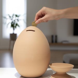 May include: A large, egg-shaped piggy bank in a neutral beige color. A hand is placing a coin into the slot on top. A small, matching pedestal dish sits beside the bank. The bank is on a white surface.