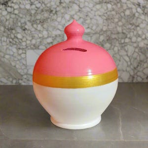 Può includere: Un salvadanaio in ceramica dipinto in tre colori. La parte superiore è rosa, separata da una fascia dorata dalla base bianca. Ha una fessura per le monete e un piccolo pinnacolo decorativo. Altezza: circa 15 cm.
