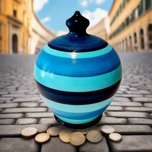 May include: A round, blue and black striped ceramic piggy bank with a black top. The bank is sitting on a cobblestone surface with several coins scattered around it. The background is a blurred image of a city street.