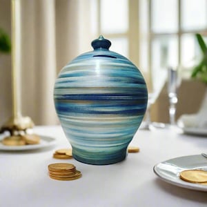 May include: A ceramic piggy bank with a blue, teal, and tan striped pattern. The bank has a small opening at the top and a dark blue finial. Gold coins are scattered around the base of the bank on a white tablecloth.