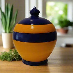 May include: A decorative, round ceramic jar with a lid. The jar is painted with alternating horizontal bands of navy blue and yellow. The jar has a dark blue base and a dark blue, pointed finial on the lid.