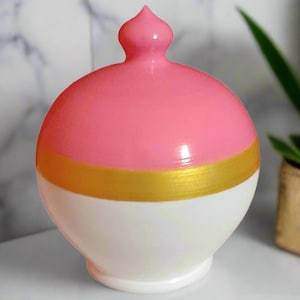 May include: A decorative ceramic jar with a rounded body and a small, pointed top. The jar is painted in three sections: a white base, a gold band, and a pink upper section. The jar is a decorative item.