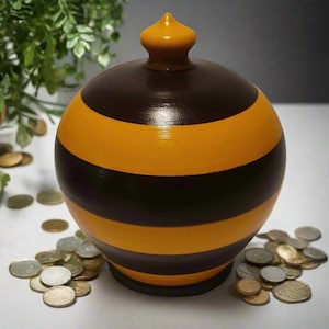 Può includere: Un salvadanaio rotondo a strisce marroni, nere e gialle, con coperchio abbinato. La banca è circondata da monete sfuse. Il design ricorda un'ape. Un oggetto decorativo per risparmiare denaro.