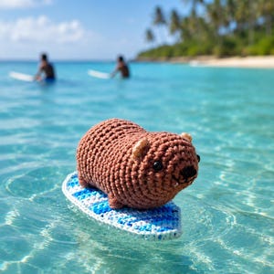 Op de afbeelding: Een bruin gehaakt capybara-figuurtje met kleine oortjes en zwarte oogjes, geplaatst op een klein, ovaalvormig surfplankje gehaakt in afwisselende tinten lichtblauw, donkerblauw en wit garen, drijvend op helder turquoise zeewater.