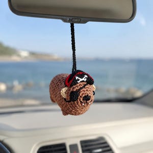 Può includere: Un ornamento da appendere a forma di capibara lavorato all'uncinetto in marrone, sospeso da un cordino nero. Il capibara presenta una benda nera sull'occhio e un cappello da pirata nero e rosso con un simbolo bianco di ossa incrociate.