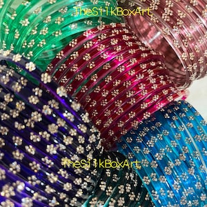 Puede incluir: Una colección de brazaletes translúcidos en verde, rosa, rojo, morado y azul, cada uno con un patrón repetitivo de pequeñas flores doradas. El texto TheSilkBoxArt es visible en color amarillo en el centro de la imagen.