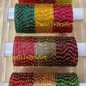May include: A collection of colorful bangles in a clear plastic container. The bangles are arranged in rows, showcasing a variety of colors including red, green, gold, and purple. The text "TheSilkBoxArt" is visible on each row.