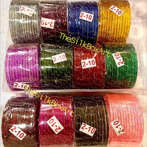 May include: A clear plastic container filled with stacks of colourful glass bangles. The bangles are in various colours, including gold, blue, red, green, and pink. Each stack has a small white tag with the number "2-10" on it.