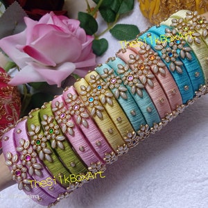 May include: A stack of colorful silk thread bangles with pearl and gemstone embellishments. The bangles are arranged in a rainbow pattern, with each bangle featuring a delicate floral design.