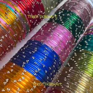 Op de afbeelding: Meerdere stapels doorschijnende glazen armbanden in diverse kleuren, waaronder rood, geel, blauw, roze, groen en goud. Elke armband is voorzien van kleine, ingebedde zilverkleurige glitteraccenten. De tekst TheSilkBoxArt is zichtbaar op de afbeelding.