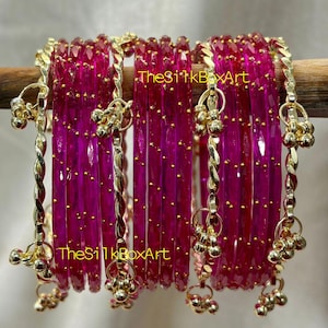 May include: A collection of magenta bangles with gold-coloured detailing. The bangles are arranged in a row and have a faceted design. Gold-coloured charms hang from the bangles. The text "TheSilkBoxArt" is visible.