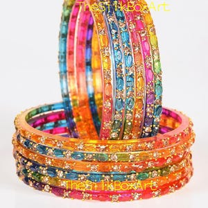 Könnte beinhalten: Ein Set aus mehrfarbigen Armreifen in leuchtenden Farben wie Pink, Orange, Gelb, Grün und Blau. Jeder Armreif ist mit einer Reihe kleiner, klarer, ovaler Steine besetzt, die in einen Metallrahmen gefasst sind. Der Text TheSilkBoxArt ist sichtbar.