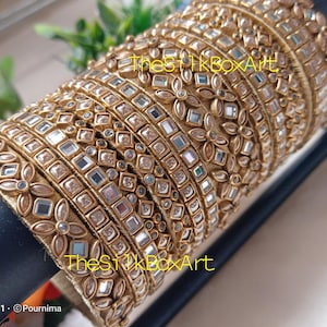 May include: A stack of gold bangles with mirrored and floral embellishments. The bangles are stacked on a black surface. The text "The Silk Box Art" is visible on the bangles.