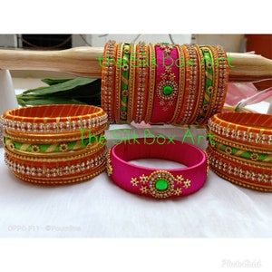 May include: A set of colorful silk thread bangles with gold and silver embellishments. The bangles are in shades of orange, green, and pink. The text "The Silk Box Art" is visible in the image.