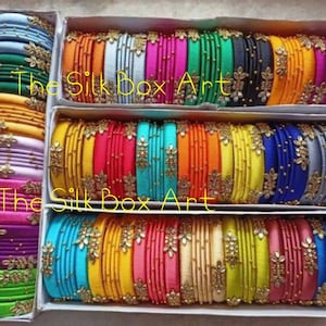 May include: A collection of colorful silk thread bangles with gold accents and pearl embellishments. The bangles are arranged in rows in white boxes. The text "The Silk Box Art" is visible on the boxes.