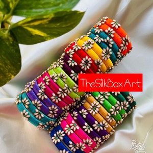 May include: A stack of colorful silk thread bangles with white flower embellishments and green gem accents. TheSilkboxArt is printed on the image.