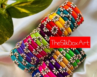 Può includere: Un mucchio di braccialetti in filo di seta colorati con decorazioni di fiori bianchi e accenti di gemme verdi. TheSilkboxArt è stampato sull'immagine.