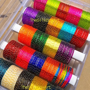 Peut inclure: Une collection de bracelets joncs multicolores disposés en rangées sur des supports cylindriques blancs à l'intérieur d'une boîte de rangement en plastique transparent. Les bracelets présentent des couleurs vives comme le rose, le vert, l'orange, le violet, le bleu, le rouge et le jaune avec des accents dorés. Le texte TheSilkBoxArt est visible sur les bracelets.