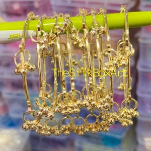 May include: A collection of gold-toned metal bangles hanging from a lime green pen. Each bangle features a thin circular band adorned with small dangling spherical charms attached by tiny rings. The text TheSilkBoxArt is visible in yellow.