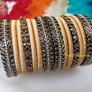 Peut inclure: Un ensemble de 12 bracelets en fil de soie dorés avec des embellissements noirs et blancs. Les bracelets sont décorés de feuilles noires et de strass blancs. Le texte "The Silk Box Art" est visible sur les bracelets.