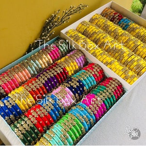 May include: A collection of colorful silk bangles with gold accents and floral designs. The bangles are arranged in a box with the text "The Silk Box Art" printed on the side.