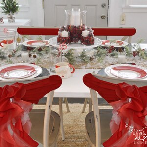 May include: A white table set for a Christmas dinner with red and white plates, red and white napkins, and a centerpiece of red berries and candles. The chairs are white with red sashes.